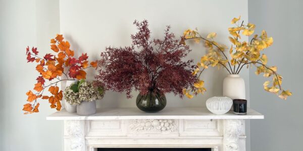 A white marble fireplace is decorated with three vases of artificial autumnal leaves. A small bunch of realistic-looking grey-green hydrangeas sits next to the vase on the far left. The vase on the left holds bright orange maple leaves on a branch. The centre vase holds a sprawling, deep burgundy-coloured branch with fine, fern-like leaves.