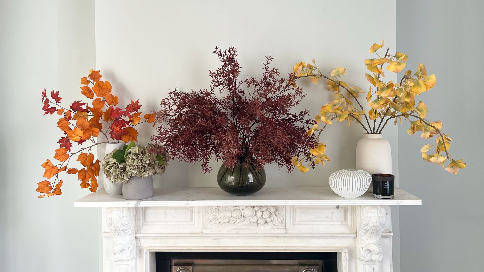 A white marble fireplace is decorated with three vases of artificial autumnal leaves. A small bunch of realistic-looking grey-green hydrangeas sits next to the vase on the far left. The vase on the left holds bright orange maple leaves on a branch. The centre vase holds a sprawling, deep burgundy-coloured branch with fine, fern-like leaves.