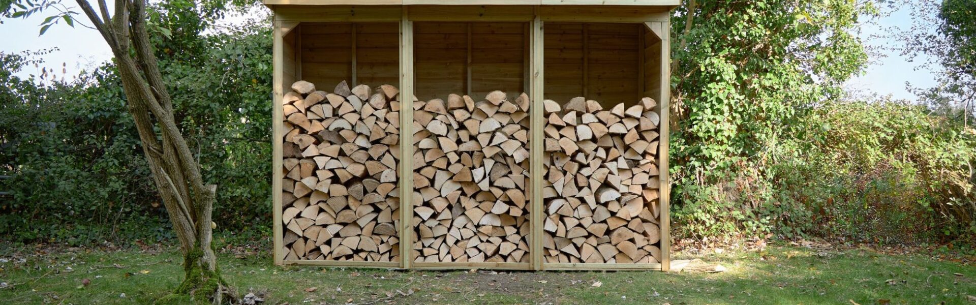 A three-bay wooden log store is filled with neatly stacked firewood. It sits on a patch of grass in front of a leafy green hedge, with trees visible on either side and in the background. The store has a sloped roof that overhangs the front.