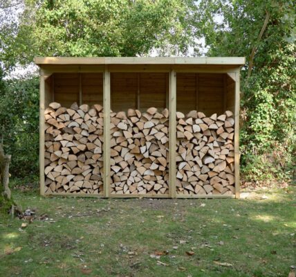 A three-bay wooden log store is filled with neatly stacked firewood. It sits on a patch of grass in front of a leafy green hedge, with trees visible on either side and in the background. The store has a sloped roof that overhangs the front.