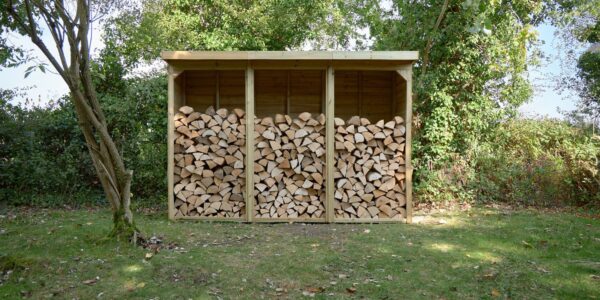 A three-bay wooden log store is filled with neatly stacked firewood. It sits on a patch of grass in front of a leafy green hedge, with trees visible on either side and in the background. The store has a sloped roof that overhangs the front.