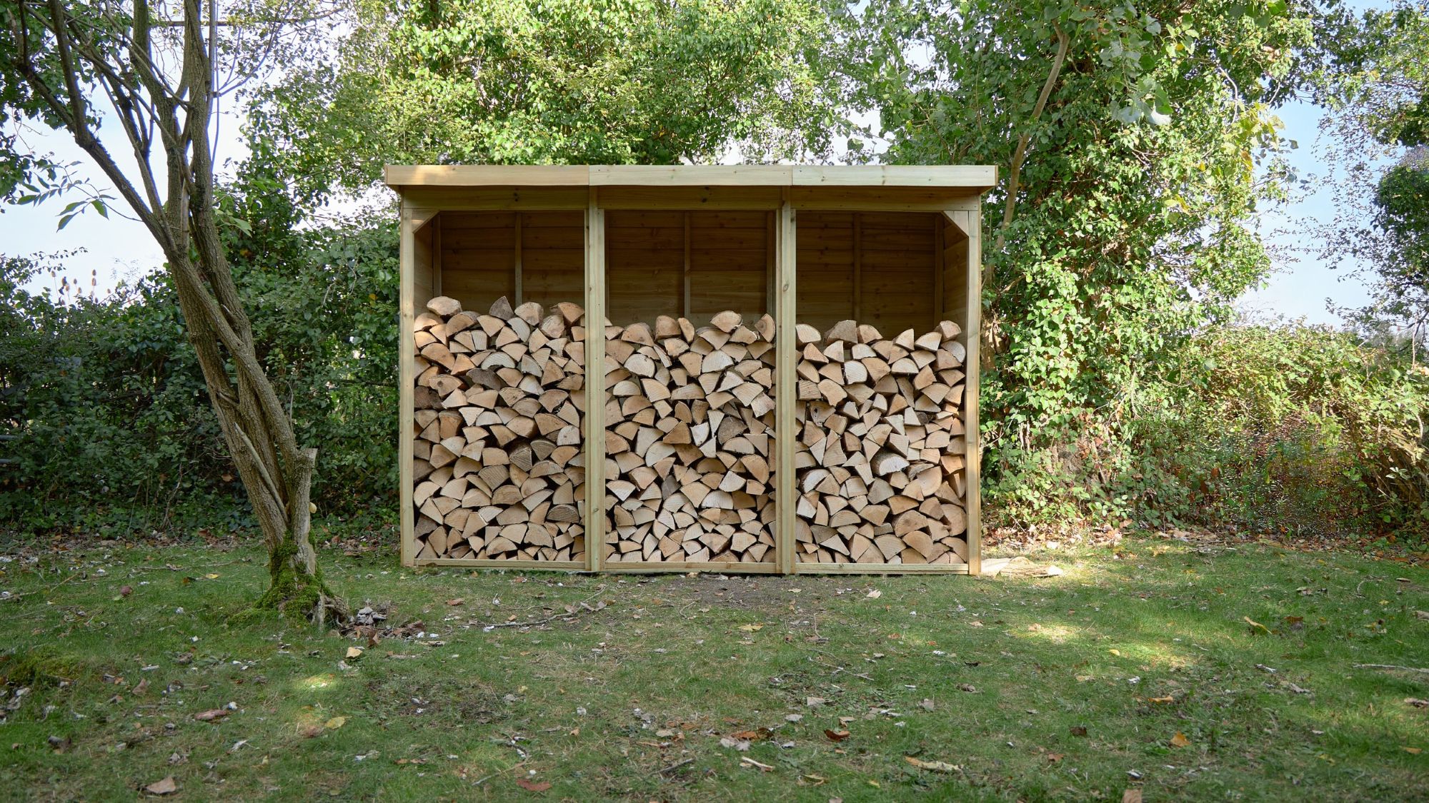 A three-bay wooden log store is filled with neatly stacked firewood. It sits on a patch of grass in front of a leafy green hedge, with trees visible on either side and in the background. The store has a sloped roof that overhangs the front.