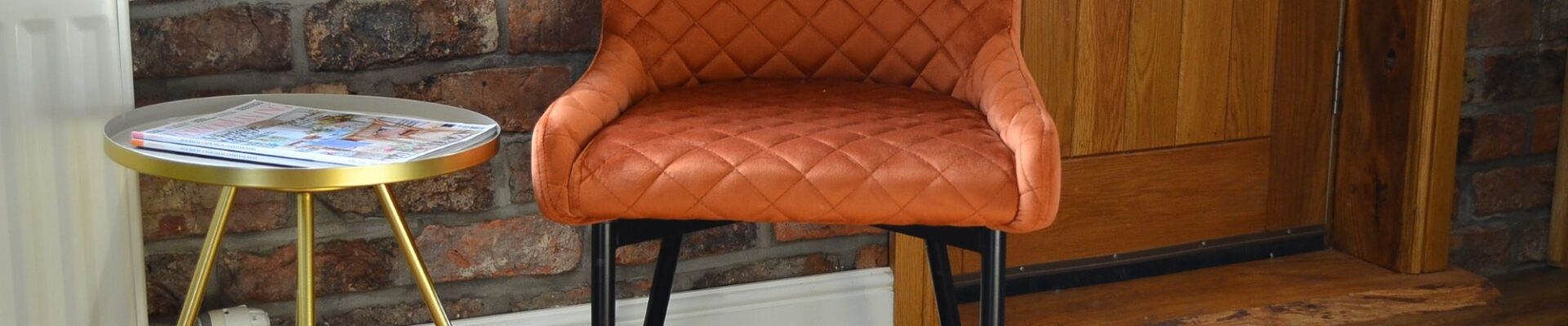 A quilted burnt orange velvet chair with black metal legs, sits near a small, round side table with a silver top and gold-coloured, tripod-style legs. A rustic brick wall behind the chair.