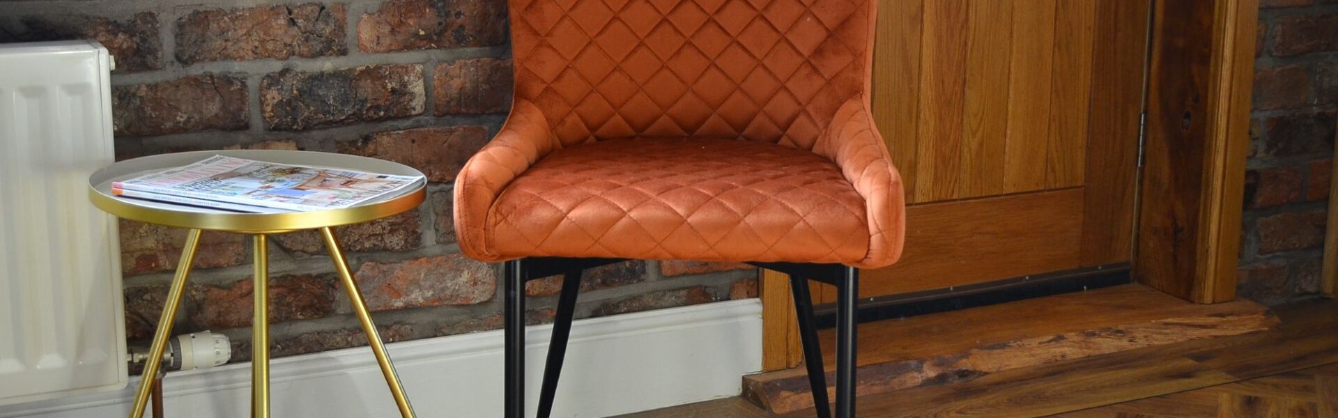 A quilted burnt orange velvet chair with black metal legs, sits near a small, round side table with a silver top and gold-coloured, tripod-style legs. A rustic brick wall behind the chair.