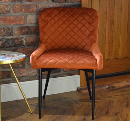 A quilted burnt orange velvet chair with black metal legs, sits near a small, round side table with a silver top and gold-coloured, tripod-style legs. A rustic brick wall behind the chair.
