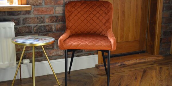 A quilted burnt orange velvet chair with black metal legs, sits near a small, round side table with a silver top and gold-coloured, tripod-style legs. A rustic brick wall behind the chair.