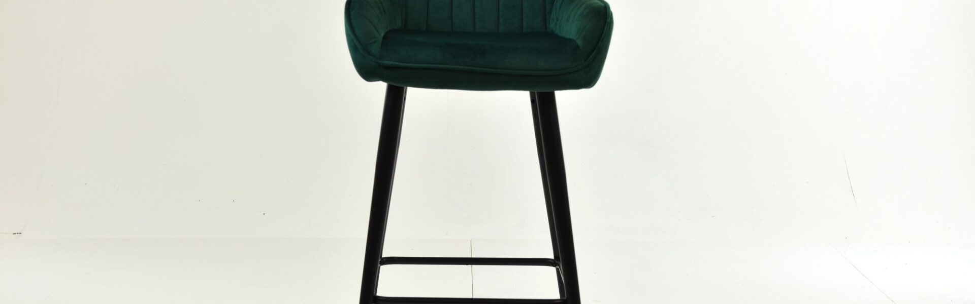 A single, modern bar stool with a deep green, vertically channel-tufted velvet seat and backrest, resting on tall, slim black wooden legs.