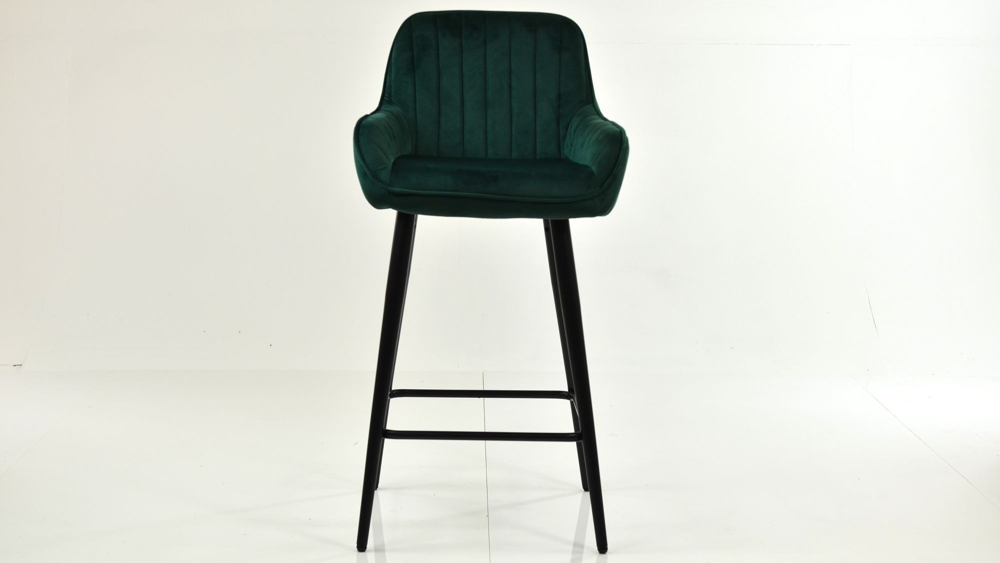 A single, modern bar stool with a deep green, vertically channel-tufted velvet seat and backrest, resting on tall, slim black wooden legs.