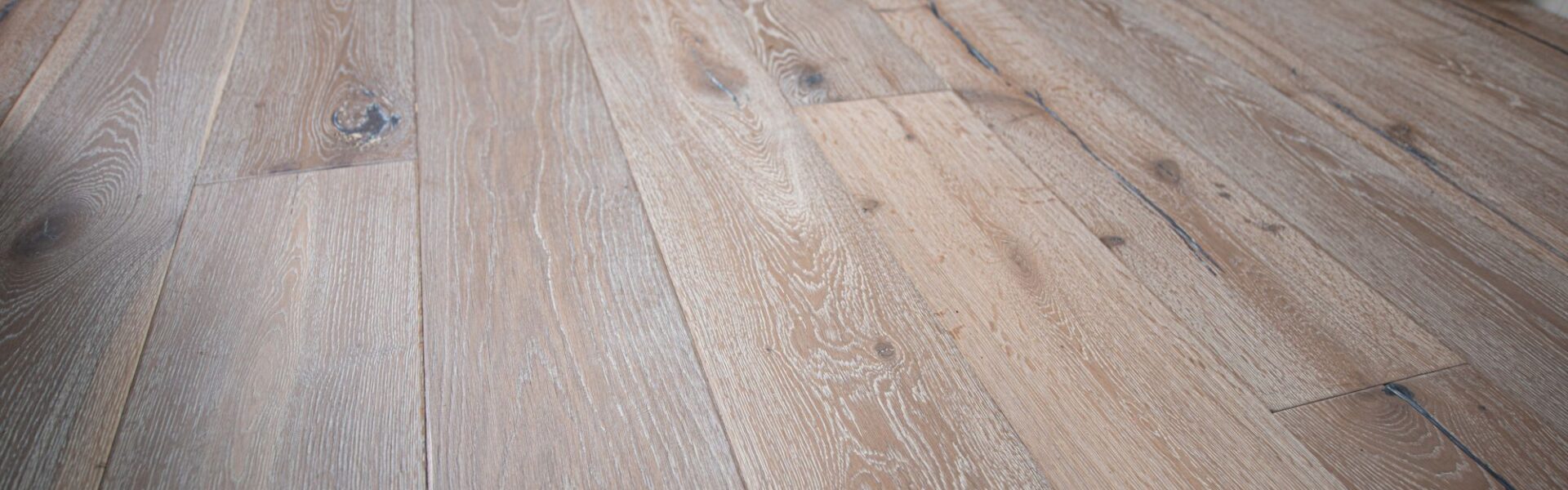 Distressed, light grey engineered oak flooring with visible wood grain and dark knots. The wide planks are laid parallel to each other, running toward a pale blue wall and a wooden cabinet. The lighting highlights the texture and varied tones of the floor.
