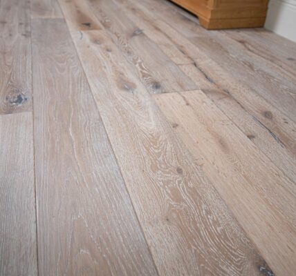 Distressed, light grey engineered oak flooring with visible wood grain and dark knots. The wide planks are laid parallel to each other, running toward a pale blue wall and a wooden cabinet. The lighting highlights the texture and varied tones of the floor.