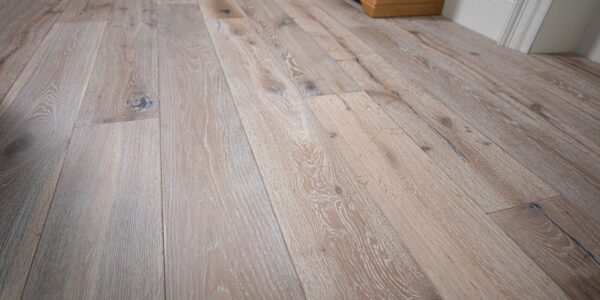 Distressed, light grey engineered oak flooring with visible wood grain and dark knots. The wide planks are laid parallel to each other, running toward a pale blue wall and a wooden cabinet. The lighting highlights the texture and varied tones of the floor.