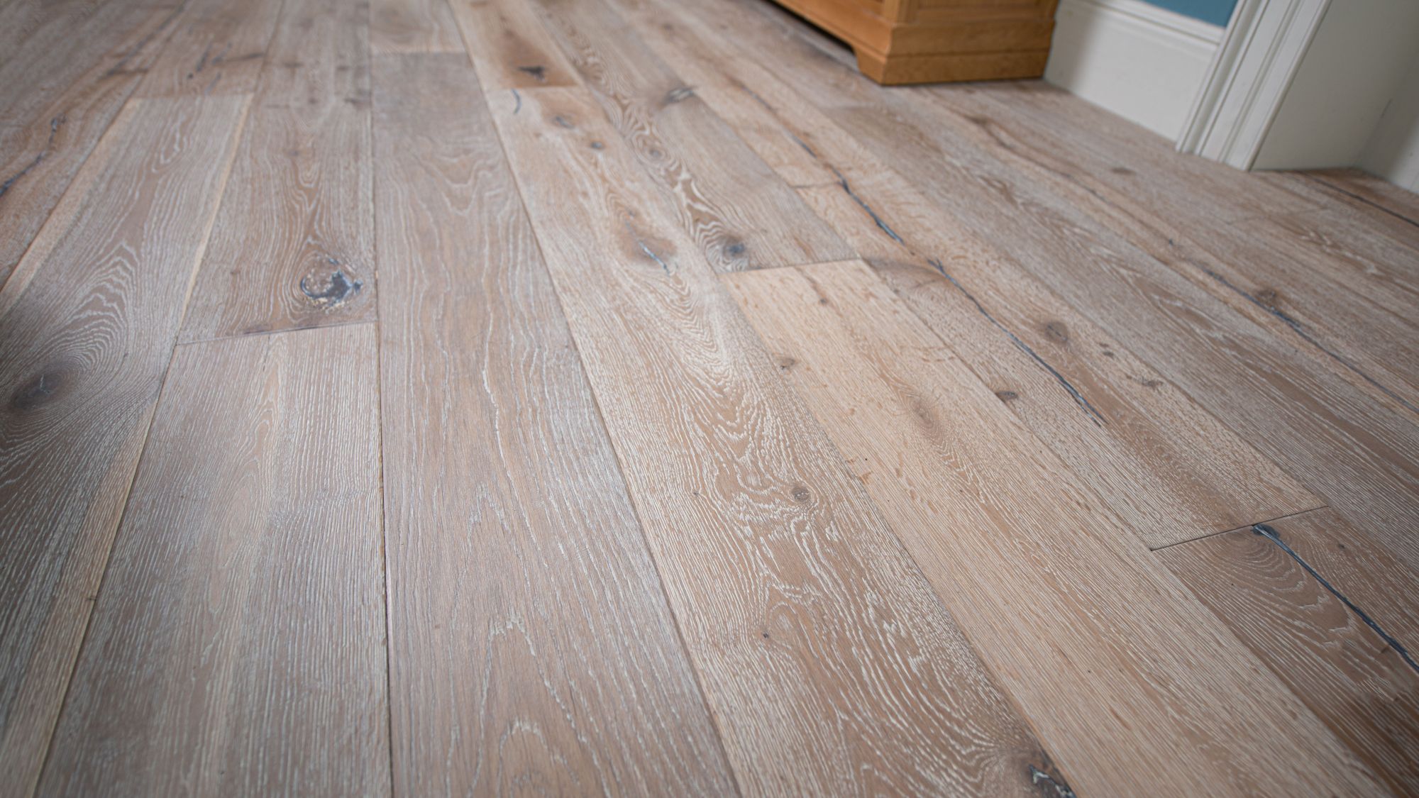 Distressed, light grey engineered oak flooring with visible wood grain and dark knots. The wide planks are laid parallel to each other, running toward a pale blue wall and a wooden cabinet. The lighting highlights the texture and varied tones of the floor.
