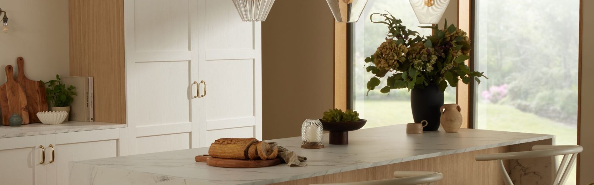 A bright kitchen island featuring a white marble countertop with grey veining and a light wood base. Two light wood and woven rush wishbone chairs are tucked underneath. Above the island, three fluted clear glass pendant lights hang.