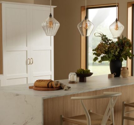 A bright kitchen island featuring a white marble countertop with grey veining and a light wood base. Two light wood and woven rush wishbone chairs are tucked underneath. Above the island, three fluted clear glass pendant lights hang.