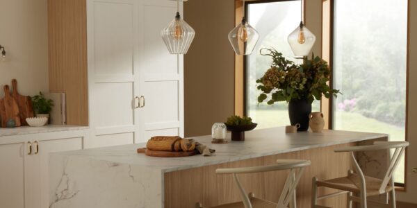 A bright kitchen island featuring a white marble countertop with grey veining and a light wood base. Two light wood and woven rush wishbone chairs are tucked underneath. Above the island, three fluted clear glass pendant lights hang.