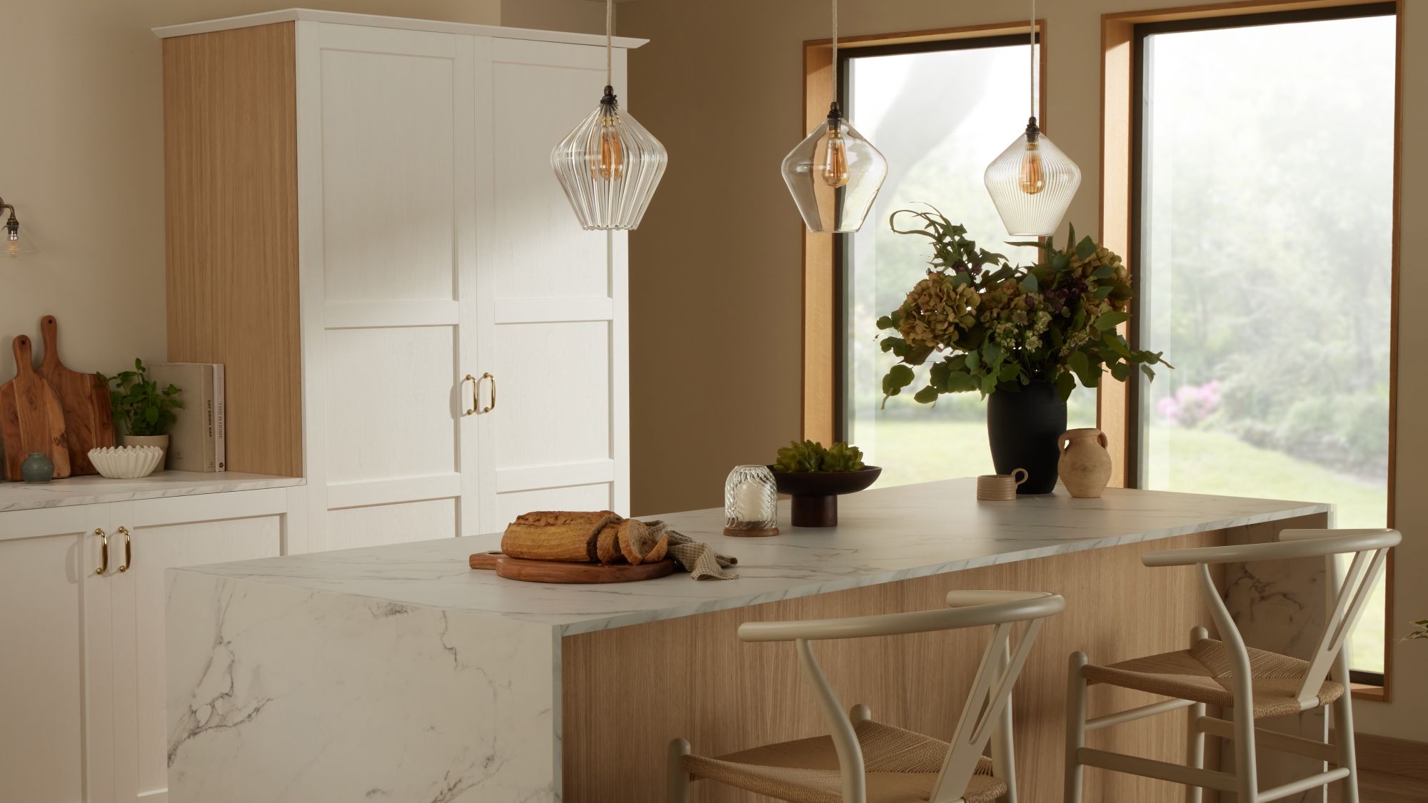 A bright kitchen island featuring a white marble countertop with grey veining and a light wood base. Two light wood and woven rush wishbone chairs are tucked underneath. Above the island, three fluted clear glass pendant lights hang.