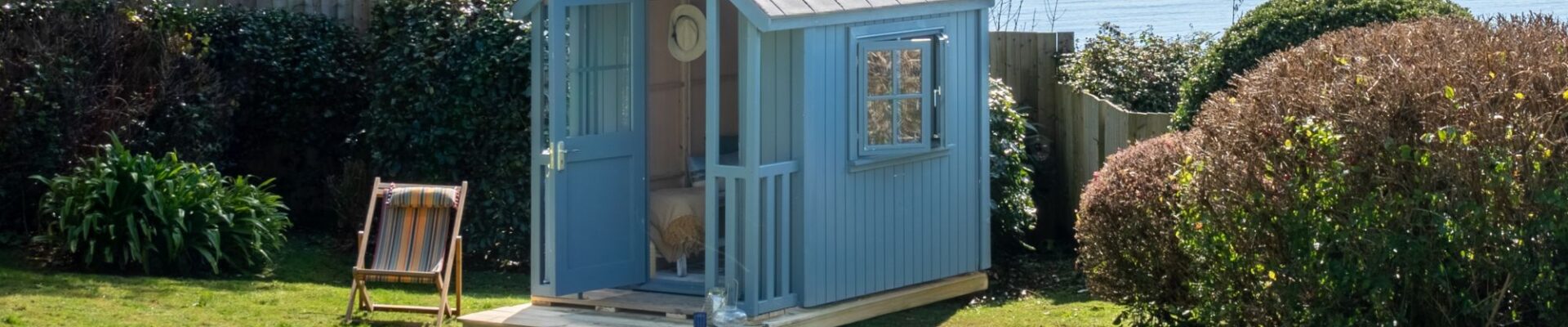 A beautiful beach hut in a garden overlooking the beach. It's painted a soft blue with a slightly darker roof and white trim around the window. It has a small porch or deck area in front of the open door.