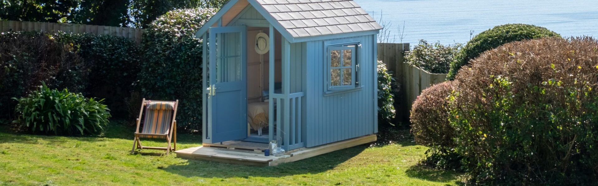 A beautiful beach hut in a garden overlooking the beach. It's painted a soft blue with a slightly darker roof and white trim around the window. It has a small porch or deck area in front of the open door.