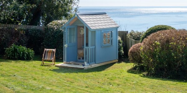 A beautiful beach hut in a garden overlooking the beach. It's painted a soft blue with a slightly darker roof and white trim around the window. It has a small porch or deck area in front of the open door.