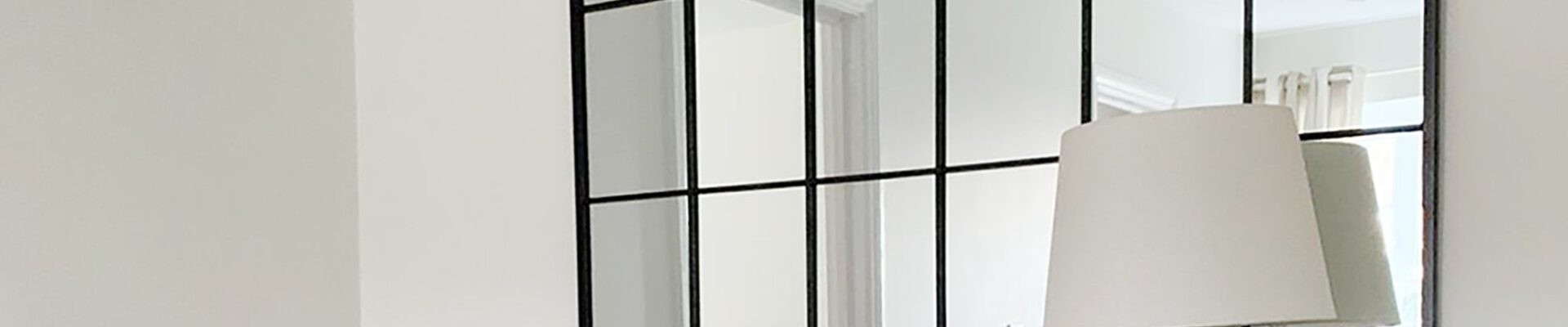 This image features a classic arch-topped, grid-style mirror with a black frame, often called a windowpane or industrial-style mirror. It's set against a very light, almost white wall. The mirror is placed above a wooden console or dresser, which holds a clear glass table lamp with a simple light-coloured shade. There's also a small framed photo next to the lamp.