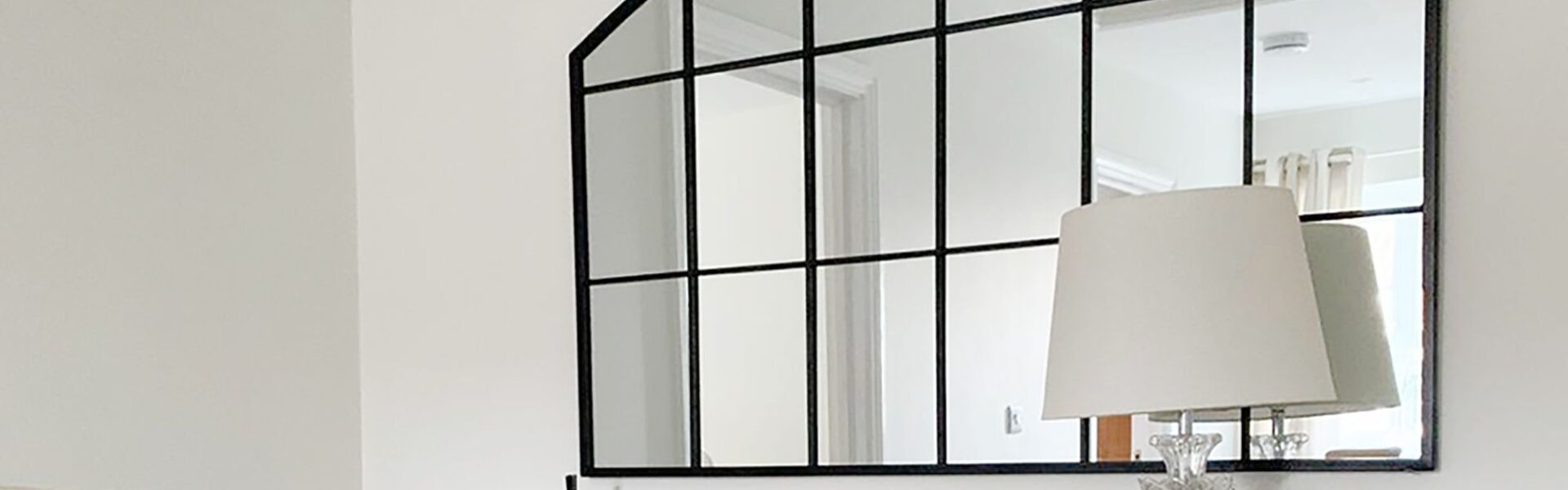 This image features a classic arch-topped, grid-style mirror with a black frame, often called a windowpane or industrial-style mirror. It's set against a very light, almost white wall. The mirror is placed above a wooden console or dresser, which holds a clear glass table lamp with a simple light-coloured shade. There's also a small framed photo next to the lamp.