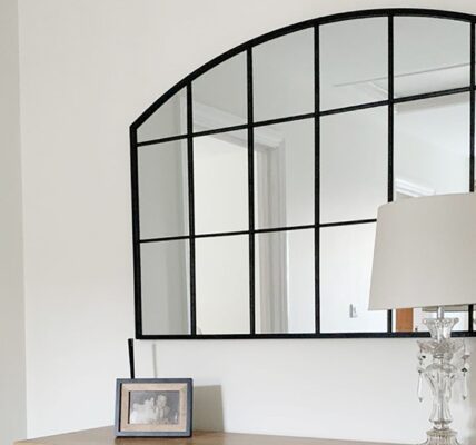 This image features a classic arch-topped, grid-style mirror with a black frame, often called a windowpane or industrial-style mirror. It's set against a very light, almost white wall. The mirror is placed above a wooden console or dresser, which holds a clear glass table lamp with a simple light-coloured shade. There's also a small framed photo next to the lamp.