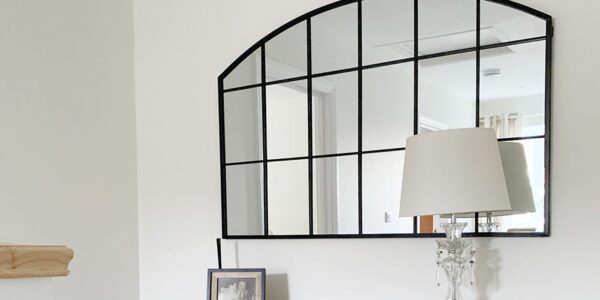 This image features a classic arch-topped, grid-style mirror with a black frame, often called a windowpane or industrial-style mirror. It's set against a very light, almost white wall. The mirror is placed above a wooden console or dresser, which holds a clear glass table lamp with a simple light-coloured shade. There's also a small framed photo next to the lamp.