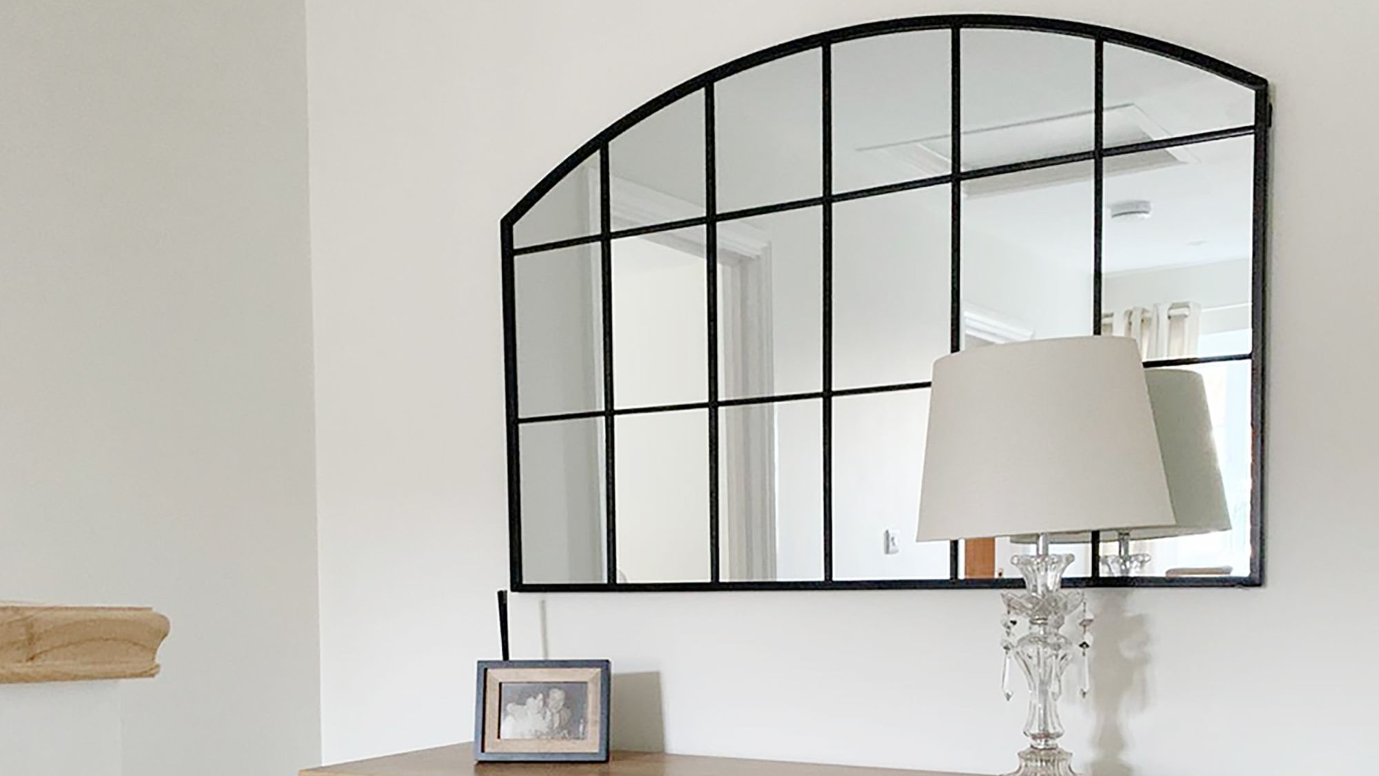 This image features a classic arch-topped, grid-style mirror with a black frame, often called a windowpane or industrial-style mirror. It's set against a very light, almost white wall. The mirror is placed above a wooden console or dresser, which holds a clear glass table lamp with a simple light-coloured shade. There's also a small framed photo next to the lamp.