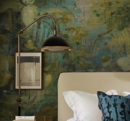 A bedroom with a textured green and gold abstract botanical wallpaper mural, featuring a bed with a beige headboard and a black-and-brass floor lamp.