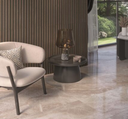 A luxurious, modern living space featuring a creamy white upholstered armchair, a ribbed wood accent wall, and glossy light-brown marble-look floor tiles. A dark round side table with a decorative lamp sits next to the chair, and a large window looks out onto a green garden.