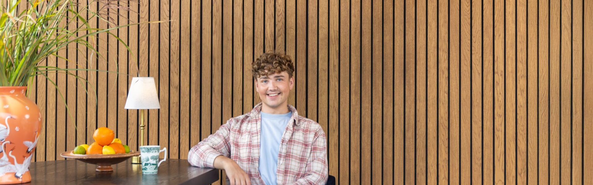 Interior Designer Jack Kinsey sits in front of a slatted wall. The slatted wall is very rich in colour and brings warmth to the image.