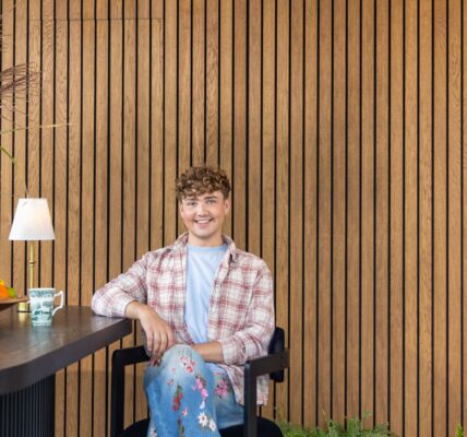 Interior Designer Jack Kinsey sits in front of a slatted wall. The slatted wall is very rich in colour and brings warmth to the image.