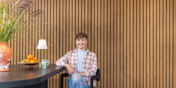 Interior Designer Jack Kinsey sits in front of a slatted wall. The slatted wall is very rich in colour and brings warmth to the image.