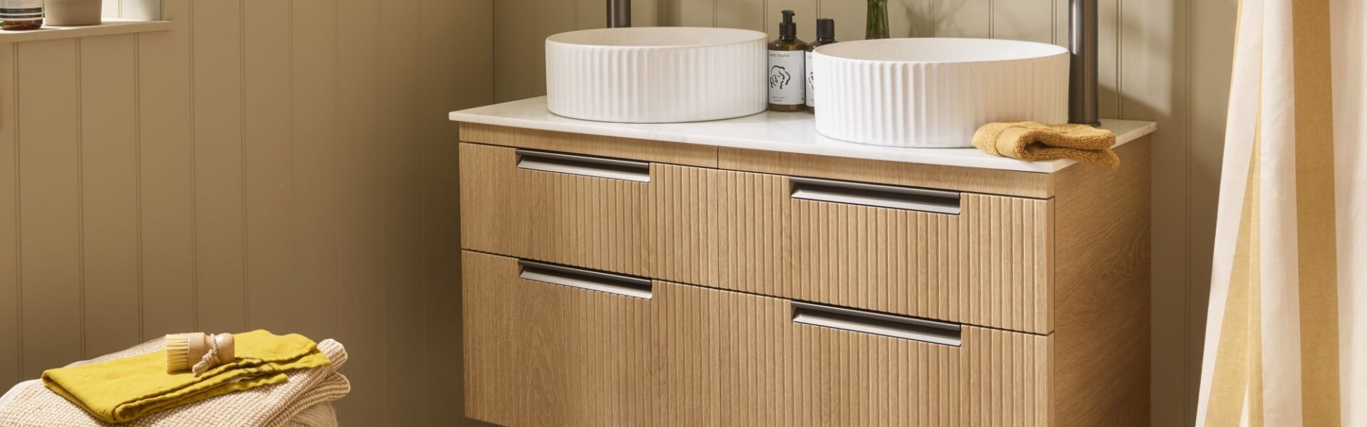 The image shows a wall-mounted vanity unit in a Swiss Oak finish with a fluted (reeded) texture on the drawer fronts. It features a double sink design with two white, ribbed/fluted countertop basins and two tall, modern gunmetal taps.