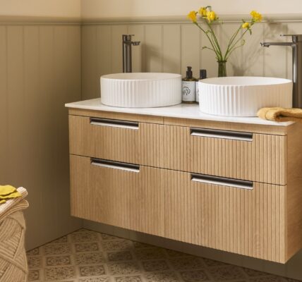The image shows a wall-mounted vanity unit in a Swiss Oak finish with a fluted (reeded) texture on the drawer fronts. It features a double sink design with two white, ribbed/fluted countertop basins and two tall, modern gunmetal taps.