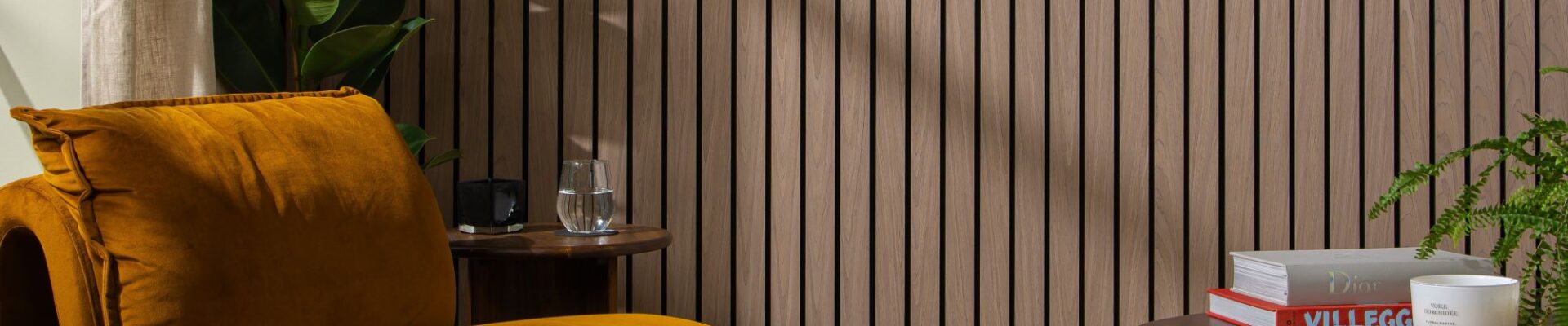 The background is dominated by a wall covered in vertical, dark walnut-coloured wooden slats set against a dark backing, creating a strong sense of texture and modern design. In the foreground on the left is a large, inviting armchair upholstered in a rich, burnt orange/mustard velvet. Its rounded, contemporary shape adds a soft contrast to the straight lines of the wall.