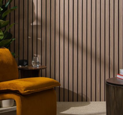The background is dominated by a wall covered in vertical, dark walnut-coloured wooden slats set against a dark backing, creating a strong sense of texture and modern design. In the foreground on the left is a large, inviting armchair upholstered in a rich, burnt orange/mustard velvet. Its rounded, contemporary shape adds a soft contrast to the straight lines of the wall.