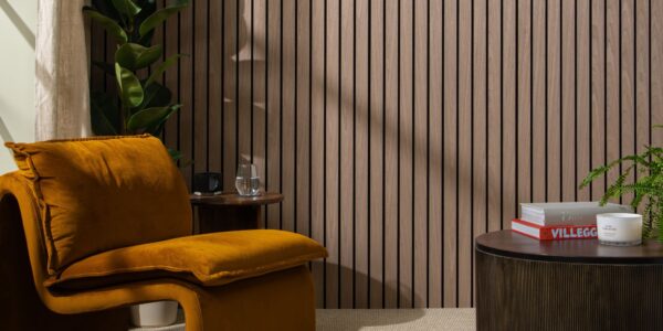 The background is dominated by a wall covered in vertical, dark walnut-coloured wooden slats set against a dark backing, creating a strong sense of texture and modern design. In the foreground on the left is a large, inviting armchair upholstered in a rich, burnt orange/mustard velvet. Its rounded, contemporary shape adds a soft contrast to the straight lines of the wall.