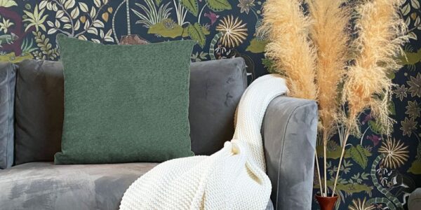 A cushion in snug, moss green colour, providing a pop of solid colour against the dark background. A sofa in a soft, dark grey or charcoal velvet/suede-like fabric which the cushion is resting on.