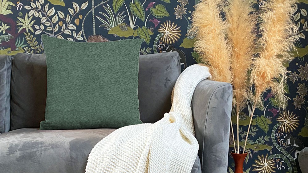 A cushion in snug, moss green colour, providing a pop of solid colour against the dark background. A sofa in a soft, dark grey or charcoal velvet/suede-like fabric which the cushion is resting on.