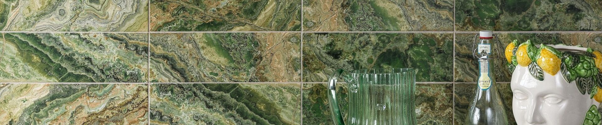 A wall tiled with rectangular, high-gloss tiles that have a dramatic, variegated pattern resembling green and brown natural stone. The dominant colours are various shades of rich green, from deep forest green to lighter lime green, mixed with earthy browns and occasional white veining, creating a strata or layered look.