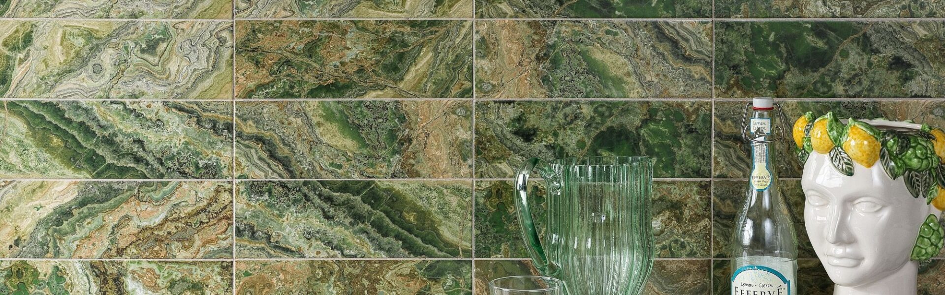 A wall tiled with rectangular, high-gloss tiles that have a dramatic, variegated pattern resembling green and brown natural stone. The dominant colours are various shades of rich green, from deep forest green to lighter lime green, mixed with earthy browns and occasional white veining, creating a strata or layered look.