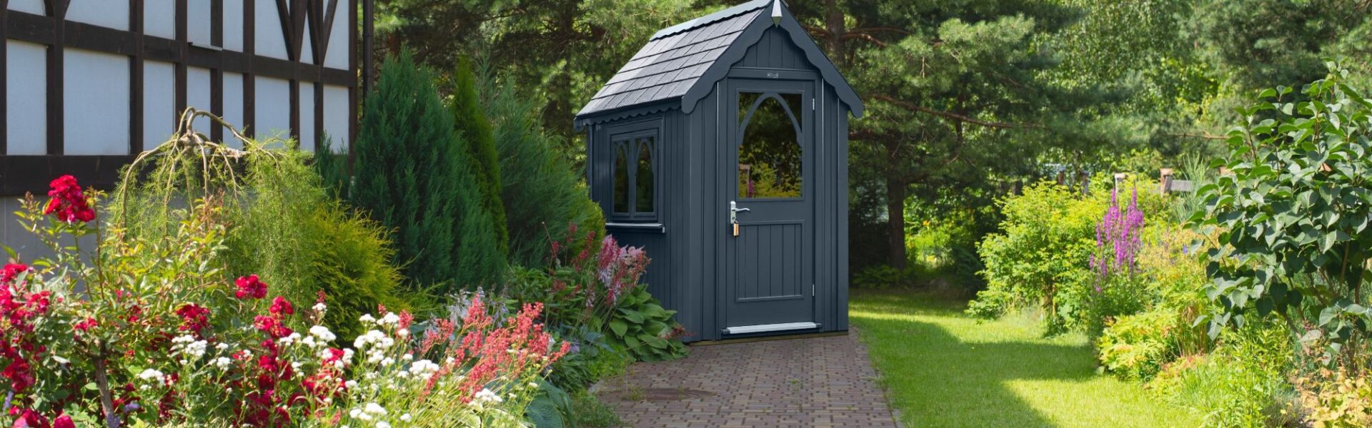 An ornate garden shed in a dark grey colour. It sits in a beautifully manicured garden.