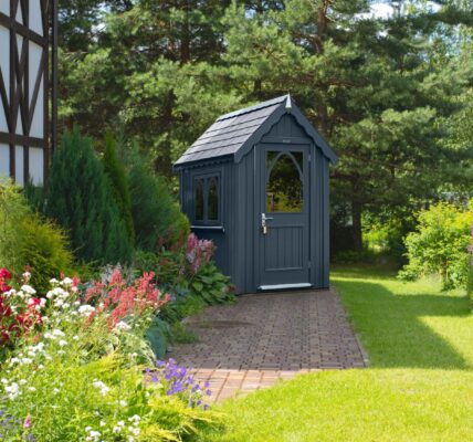 An ornate garden shed in a dark grey colour. It sits in a beautifully manicured garden.