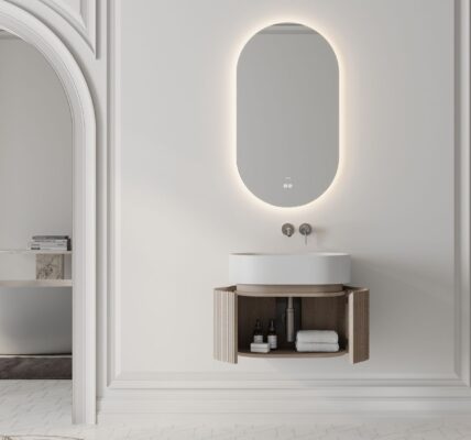A modern, minimalist bathroom design. The central focus is the vanity area, which features: a wall-mounted, oval, LED-backlit mirror, a floating vanity unit with a curved, natural wood finish and open storage, a white, counter-top basin bowl and wall-mounted taps.