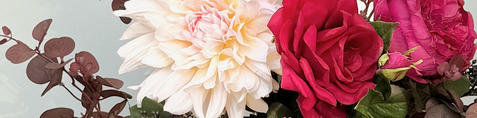 A close-up of an artificial flower bouquet featuring a large, creamy white dahlia, a deep red rose, and a magenta peony, surrounded by burgundy eucalyptus leaves and small dark berries.
