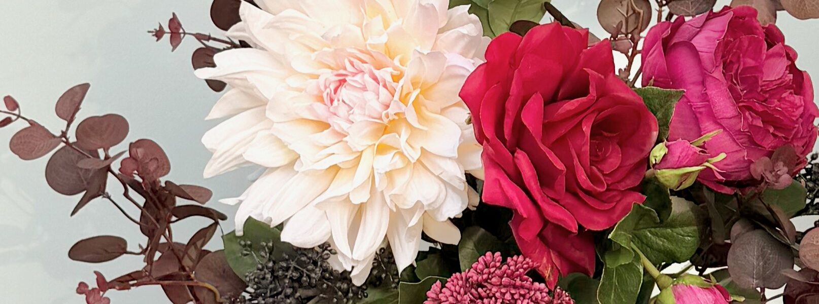 A close-up of an artificial flower bouquet featuring a large, creamy white dahlia, a deep red rose, and a magenta peony, surrounded by burgundy eucalyptus leaves and small dark berries.