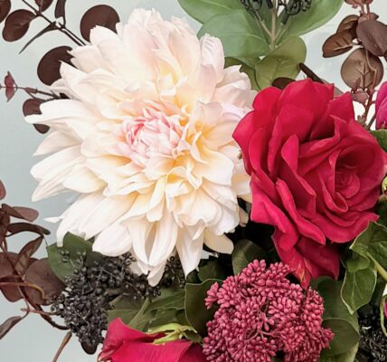 A close-up of an artificial flower bouquet featuring a large, creamy white dahlia, a deep red rose, and a magenta peony, surrounded by burgundy eucalyptus leaves and small dark berries.