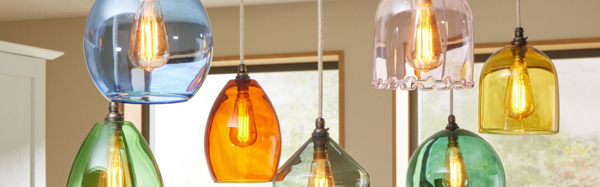 The image shows a cluster of seven colourful glass pendant lights hanging from the ceiling, likely over a kitchen island or dining area. Each pendant has a distinct shape and colour, creating a vibrant, eclectic look.