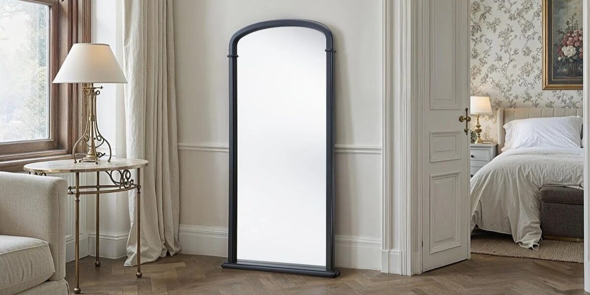 A beautiful full-length mirror with an arched top, set in a black frame. It appears to be leaning against a wall in what looks like a bedroom or dressing area. The room has classic features like hardwood floors, white trim, and patterned wallpaper. A luxurious-looking bed is visible through an open doorway, and a side table with a lamp is in the foreground.